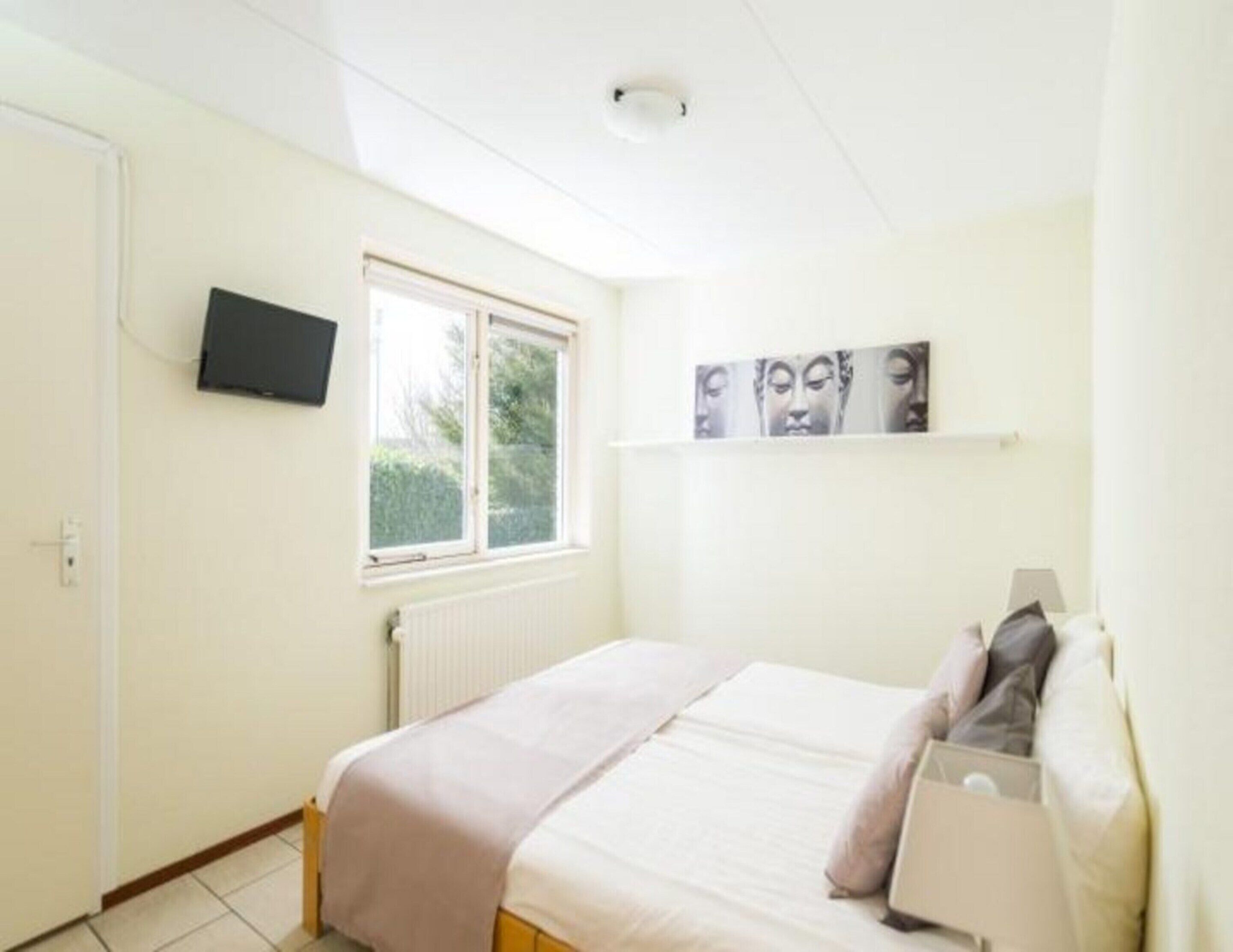 A bedroom with a bed, a window, a wall-mounted TV, and a painting.