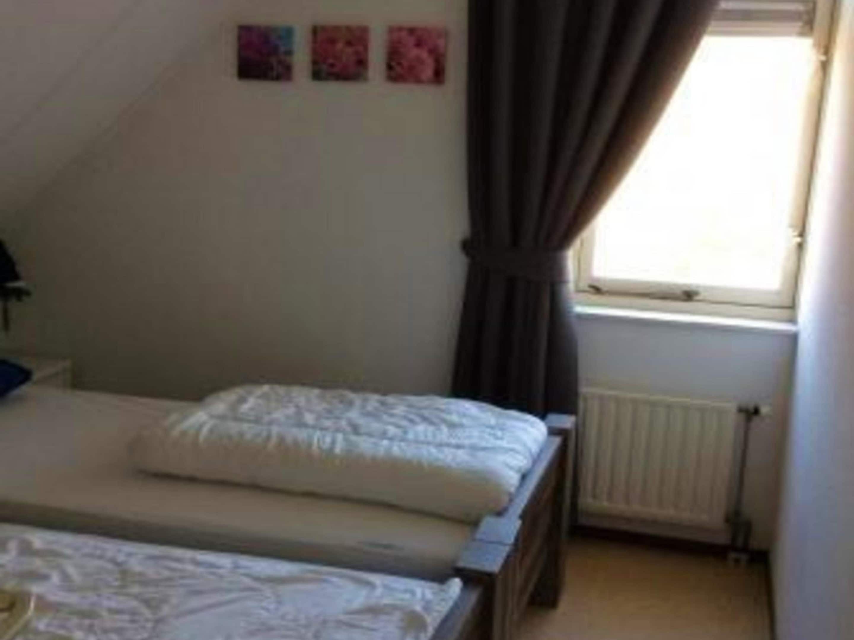 A bedroom with a bed, a window with curtains, a radiator, and three small pictures on the wall.