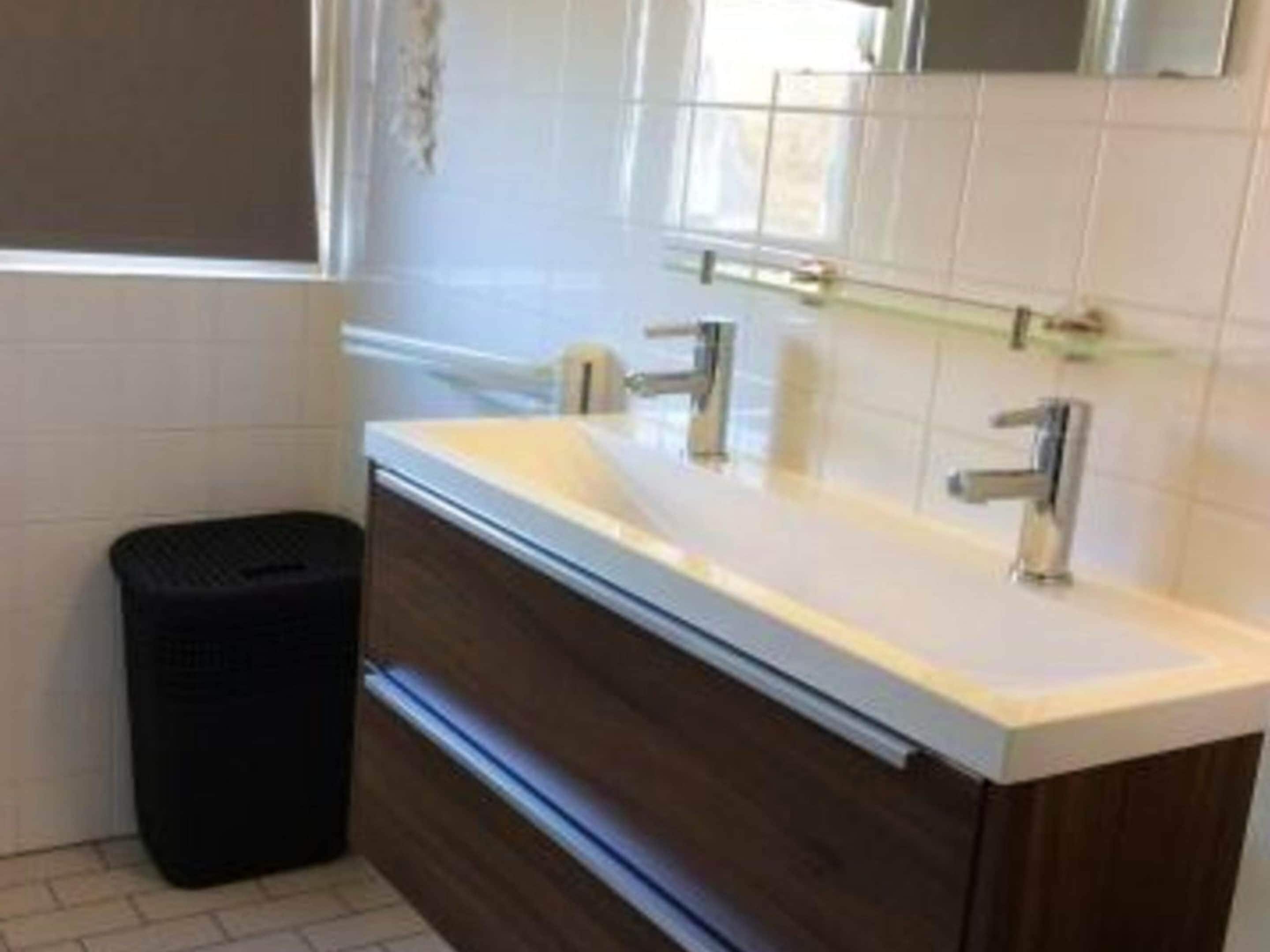 Une salle de bain avec un lavabo blanc, deux robinets, un miroir et une poubelle.