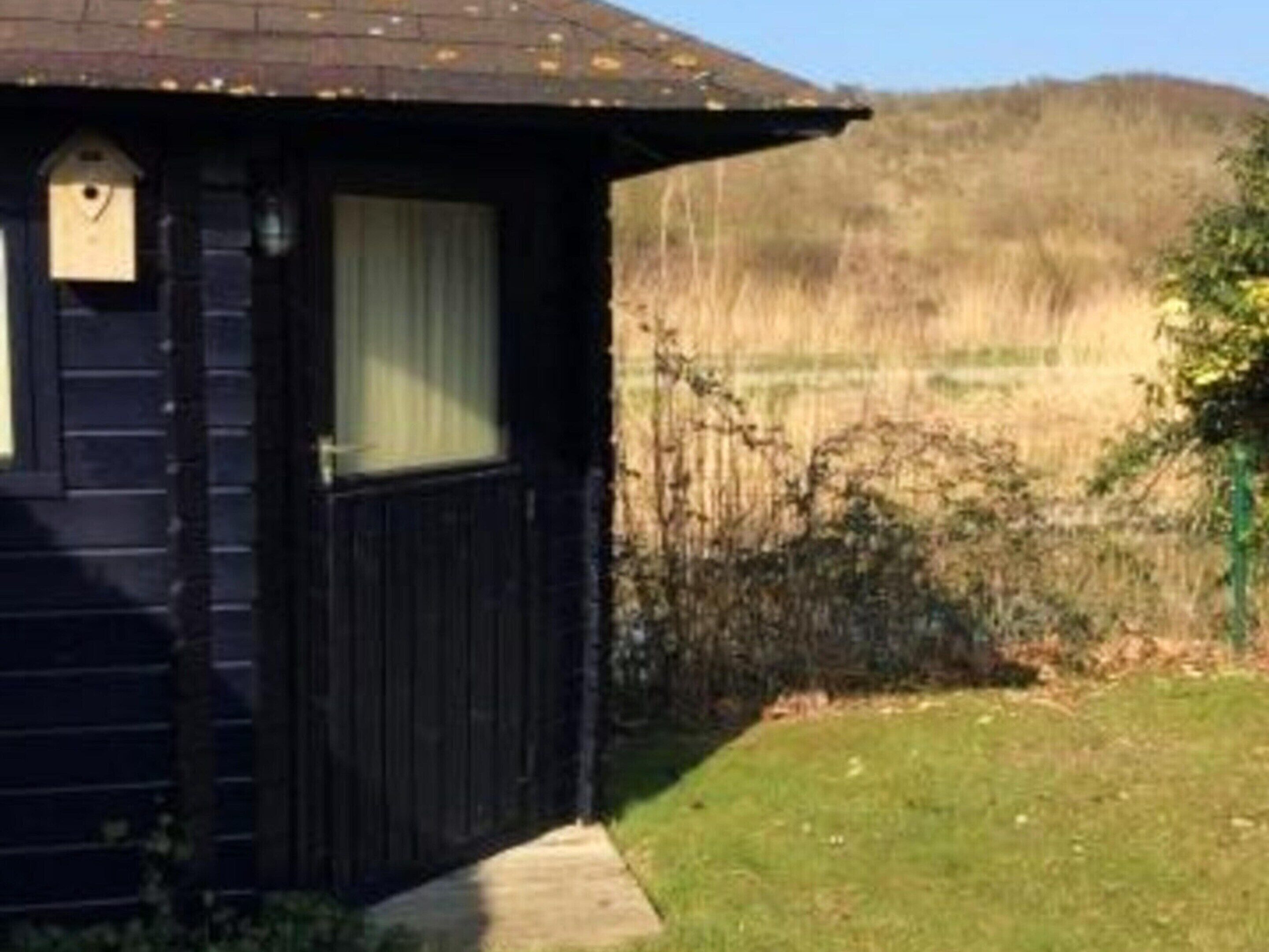 A small wooden cabin with a window and a door, surrounded by a grassy area and a fence.
