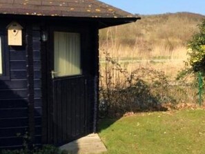 Een klein houten huisje met raam en deur, omringd door gras en een hek.