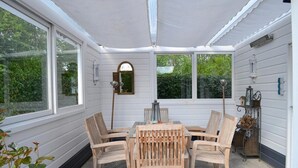 Eine überdachte Terrasse mit Tisch und Stühlen, großen Fenstern und dekorativen Wandleuchten.