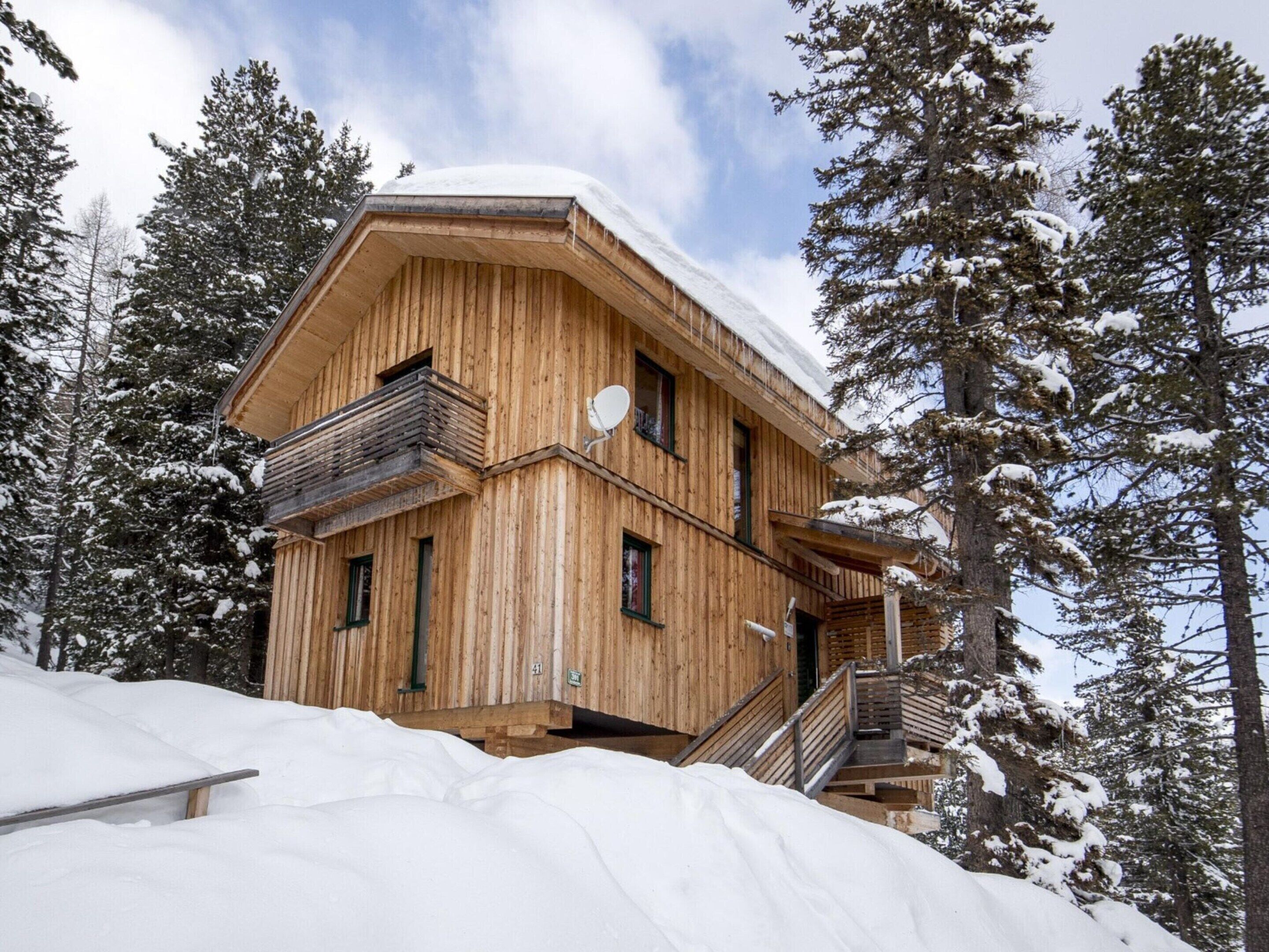A wooden chalet with a balcony and a satellite dish, surrounded by snow-covered trees.