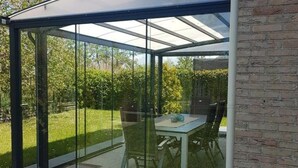 A modern glass-enclosed patio with a table and chairs, surrounded by a brick wall and a well-maintained garden. - Coastal Bungalow w/ Garden (Breskens)