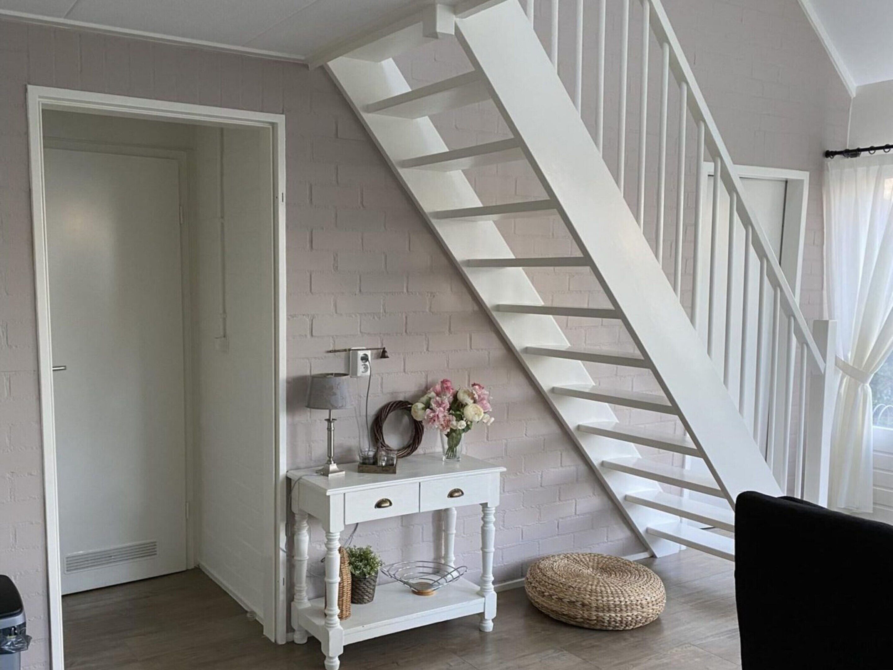 A staircase with white railings, a white console table with drawers, a wicker basket, and a small round ottoman.