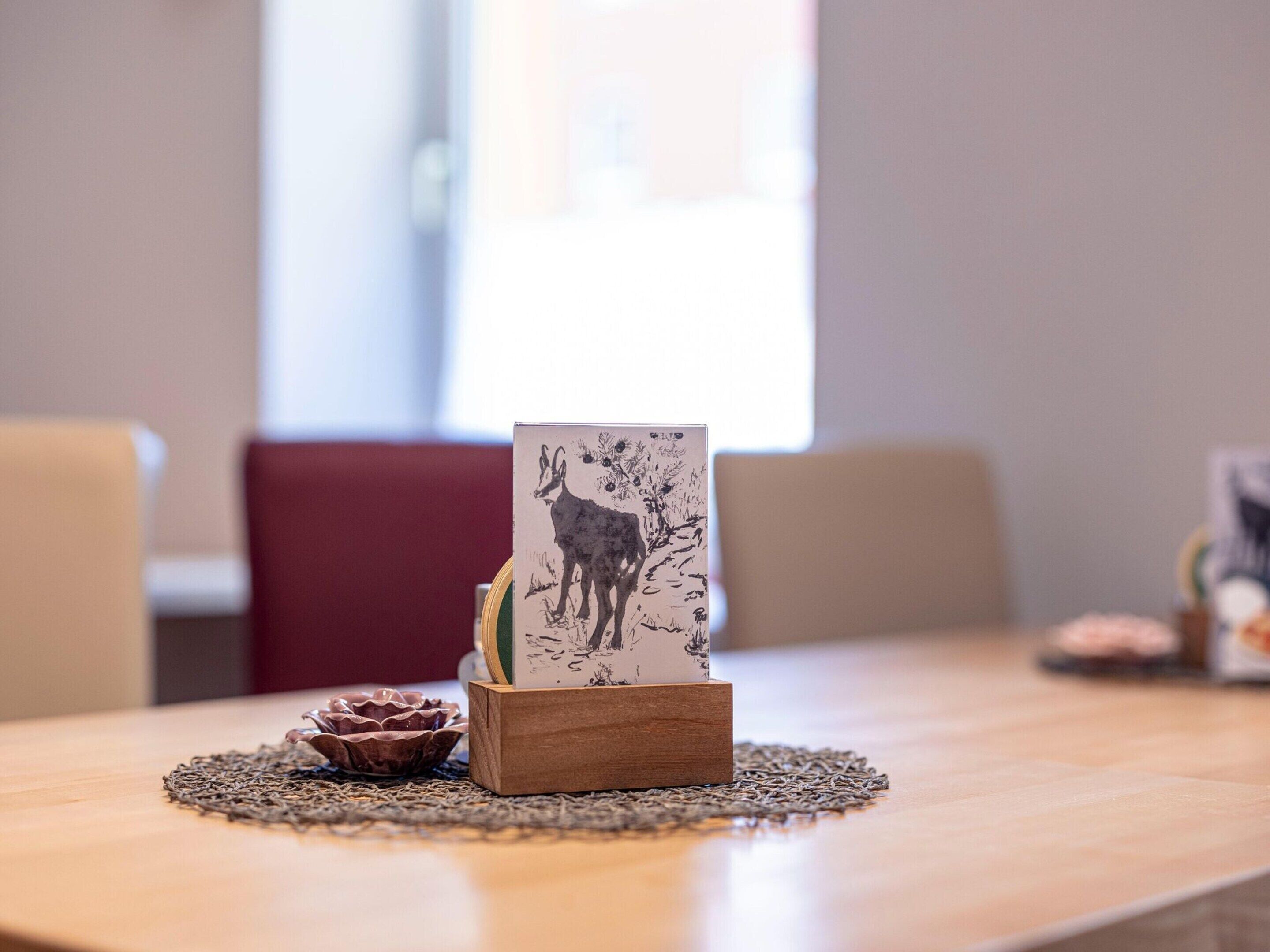 Una mesa de madera con un mantel individual, un pequeño bloque de madera con una ilustración blanco y negro de un ciervo, y un tazón decorativo.