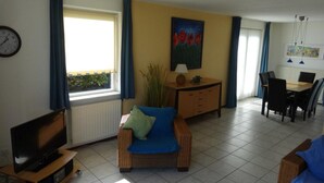 A living room with a sofa, a TV on a stand, a dining table with chairs, a painting on the wall, and a window with curtains. - Bungalow in Breskens Near the Lighthouse (Breskens)