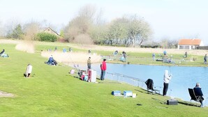 Eine Seenlandschaft mit Menschen beim Angeln, einer grünen Wiese und einem Haus im Hintergrund.