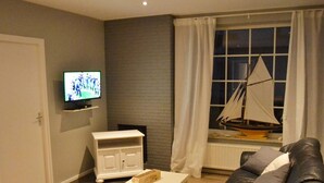 Ein Wohnzimmer mit Fernseher, einem kleinen Schrank, einem Tisch, einer Couch und einem Schiffsmodell am Fenster.