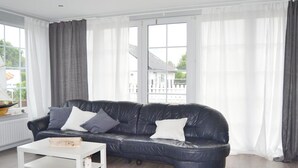 Ein modernes Wohnzimmer mit einer schwarzen Ledersofa, weißen Vorhängen und Blick auf ein Gebäude.