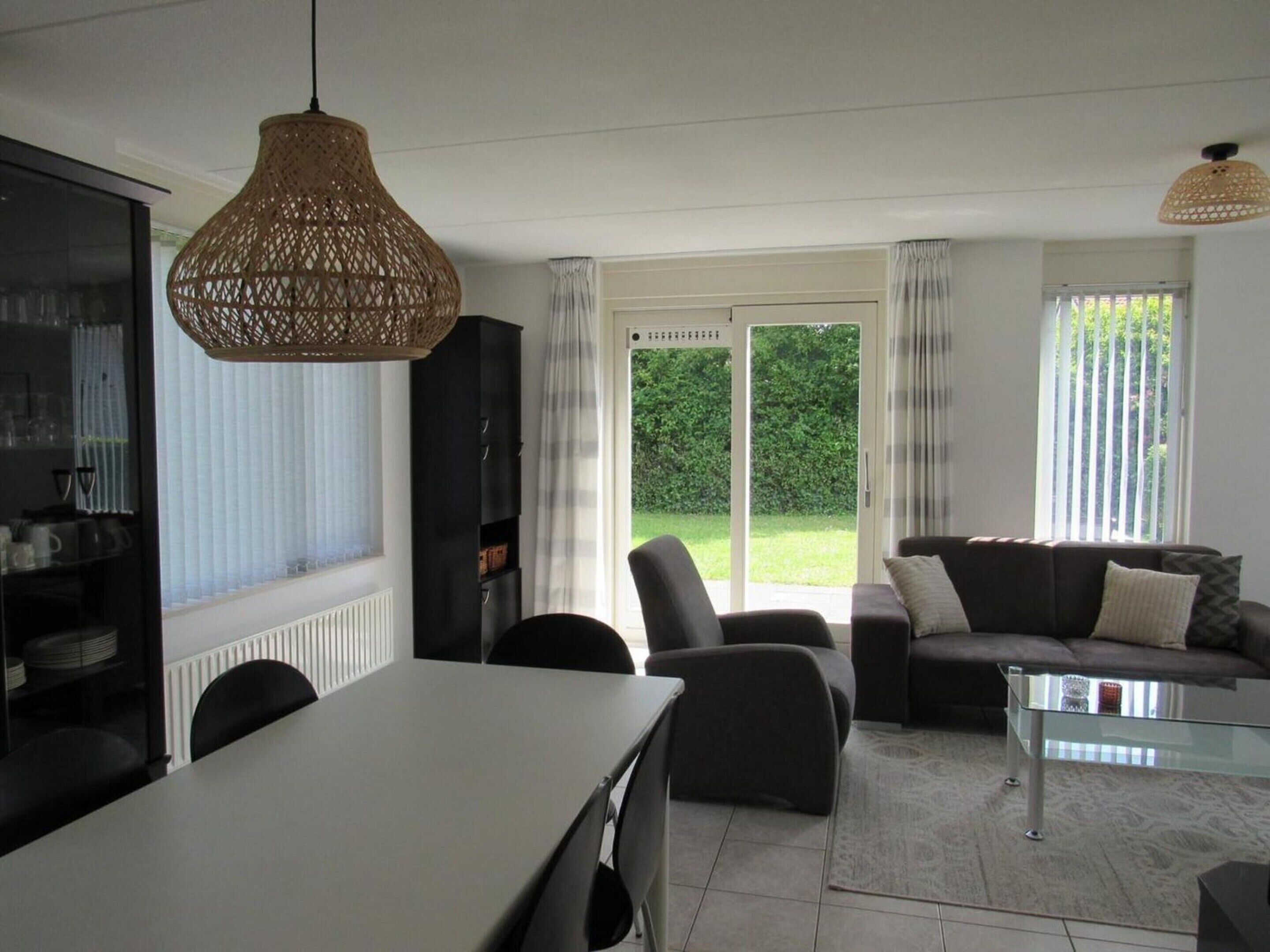 A modern living room with a dining table, chairs, a sofa, and a glass coffee table. There is a large window with a view of a green garden.