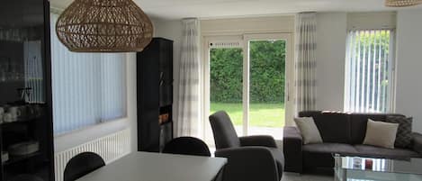 A modern living room with a dining table, chairs, a sofa, and a glass coffee table. There is a large window with a view of a green garden.