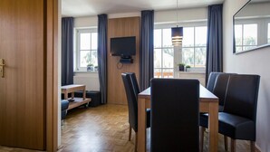 A modern room with a wooden dining table, black chairs, a flat-screen TV mounted on the wall, and large windows with curtains. - Studio Apartment in Alps Near Ski Station (Stadl-Predlitz)