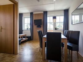 A modern room with a wooden dining table, black chairs, a flat-screen TV mounted on the wall, and large windows with curtains.