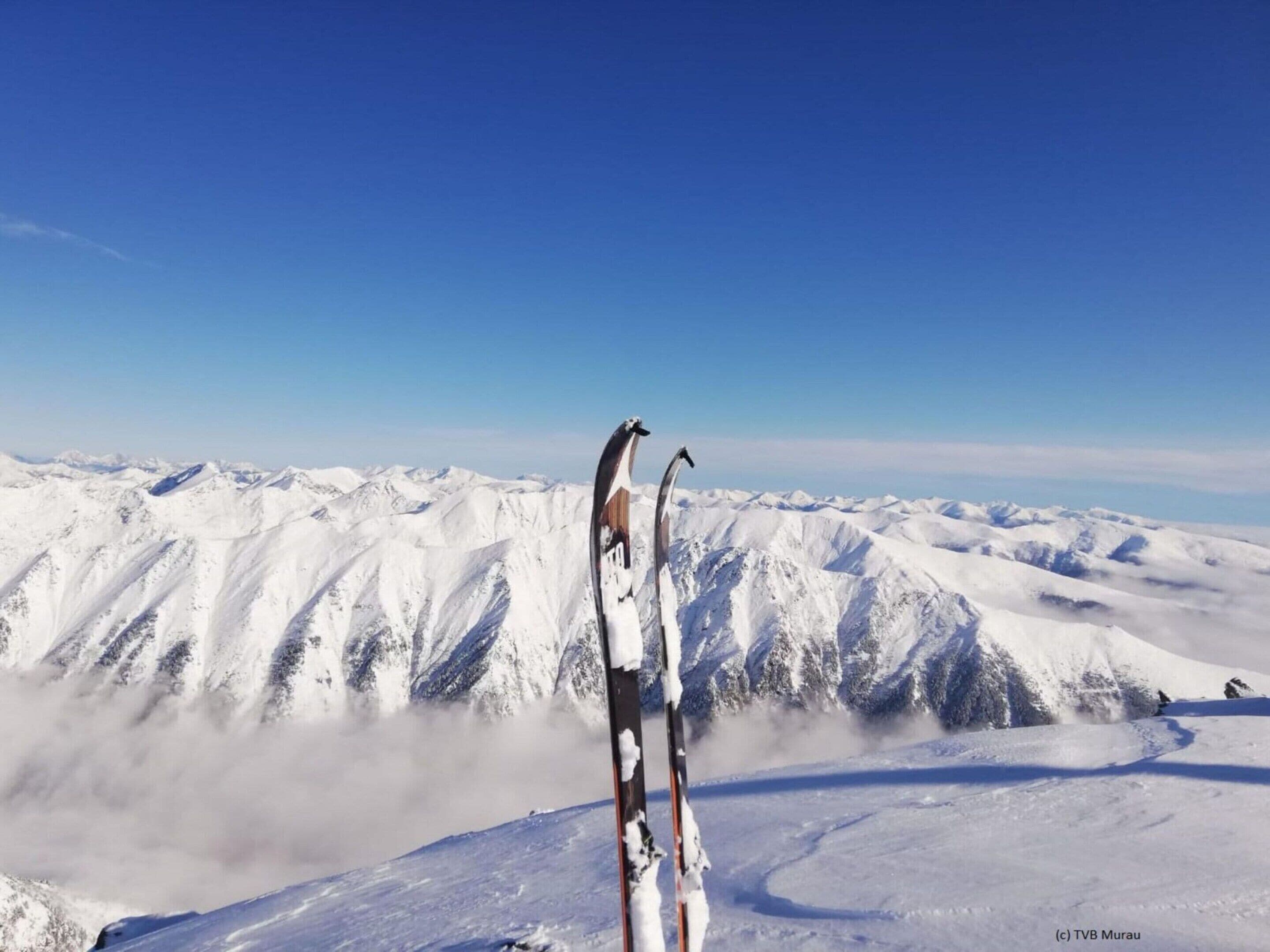 A pair of skis standing upright on a snow-covered mountain peak with a clear blue sky in the background.