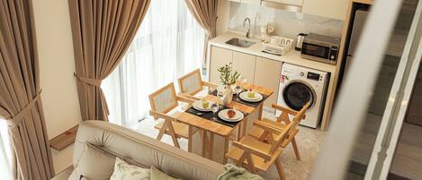 A modern living room with a sofa, dining table, and chairs, a kitchen with appliances, and large windows with curtains.
