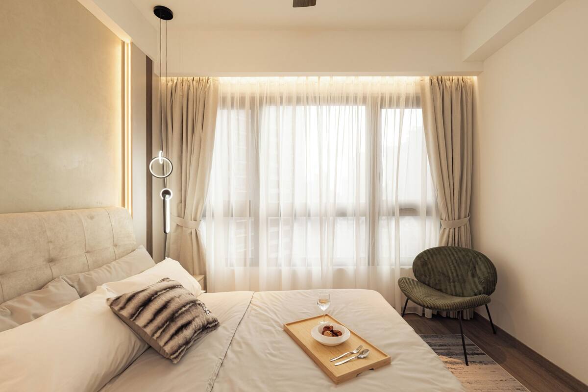 Una habitación de hotel moderna con una cama grande, una mesita de noche con una bandeja de desayuno, una silla y cortinas de gasa.