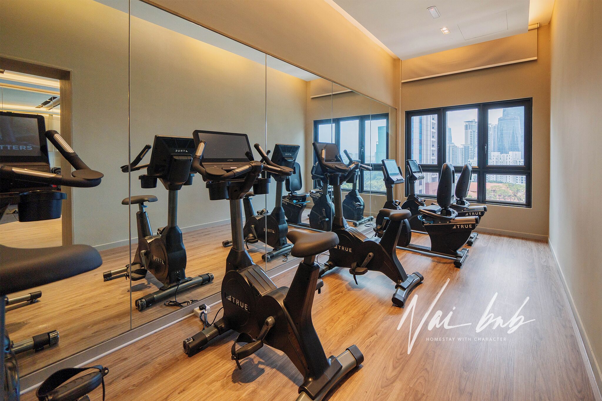 A gym with multiple exercise bikes and a mirrored wall, large windows with a city view, and a wooden floor.