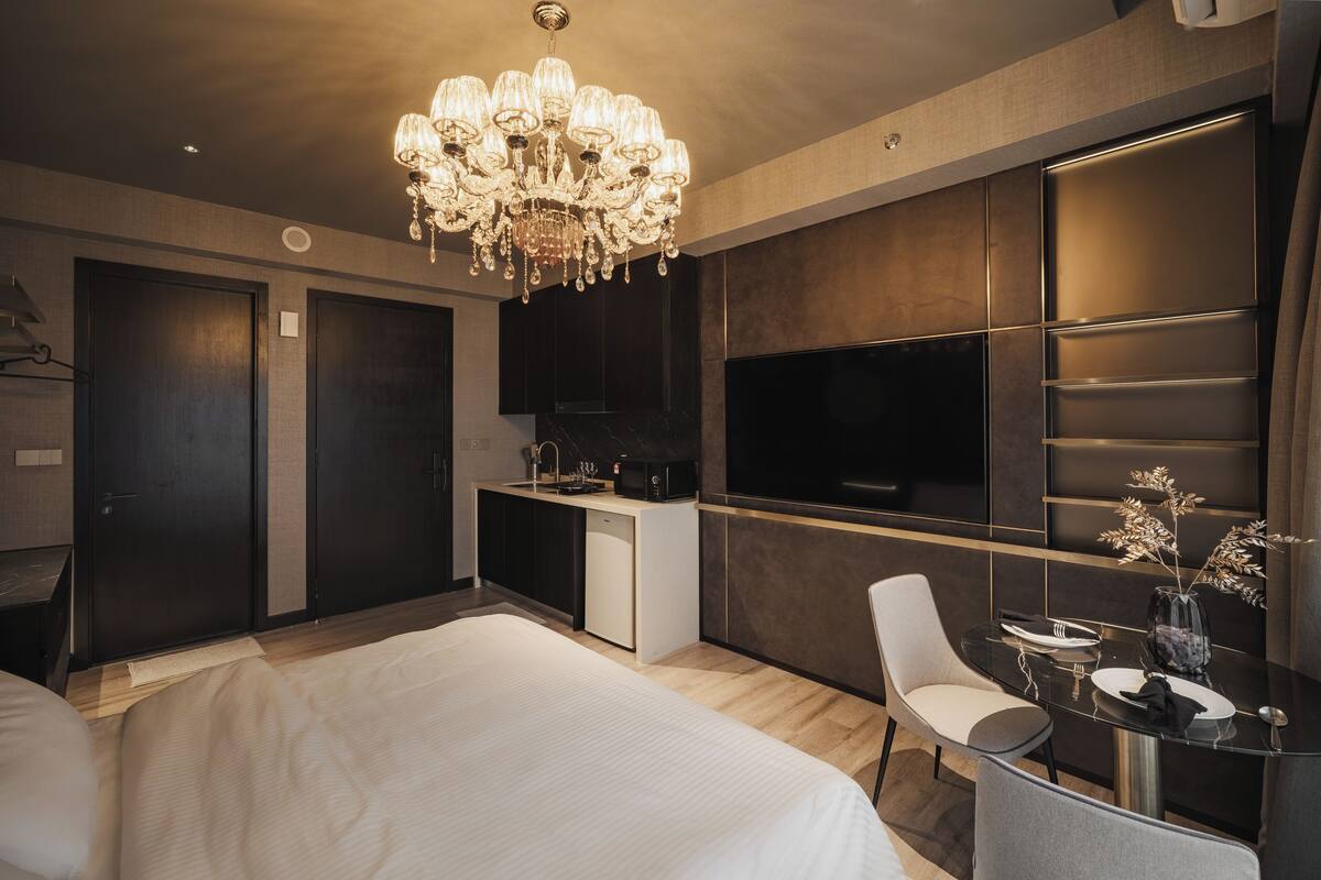 Habitación de hotel moderna con una cama grande, una mesa de comedor con sillas, un candelabro, un televisor de pantalla plana y un minibar.