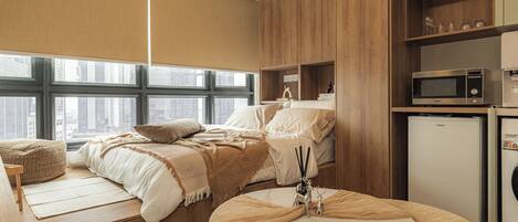 A compact hotel room with a bed, seating area, kitchenette, and a view of the city.