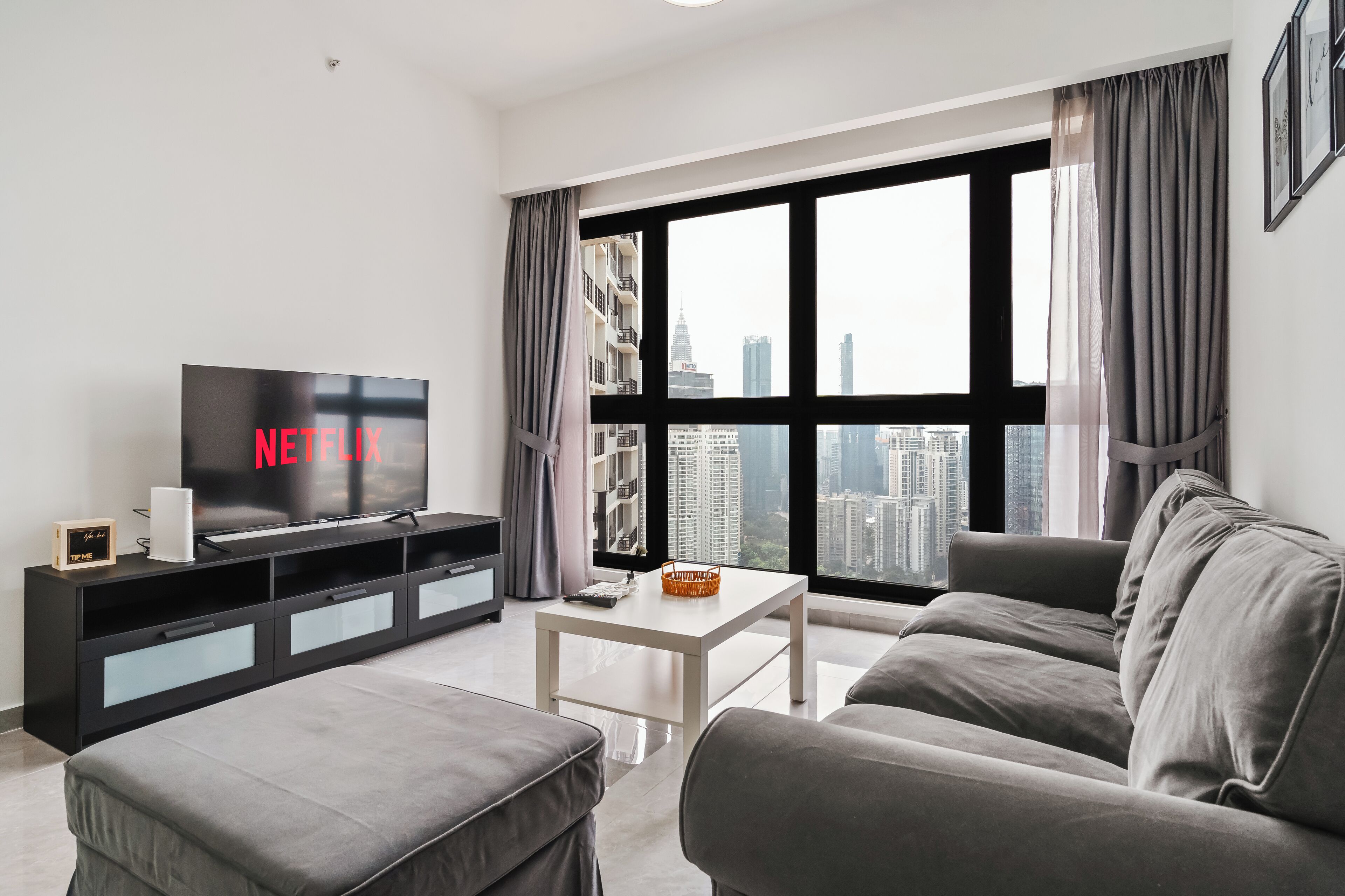 Een moderne woonkamer met een grote televisie waarop “NETFLIX” staat, een bank, een salontafel en een uitzicht op de stadslijn door grote ramen.