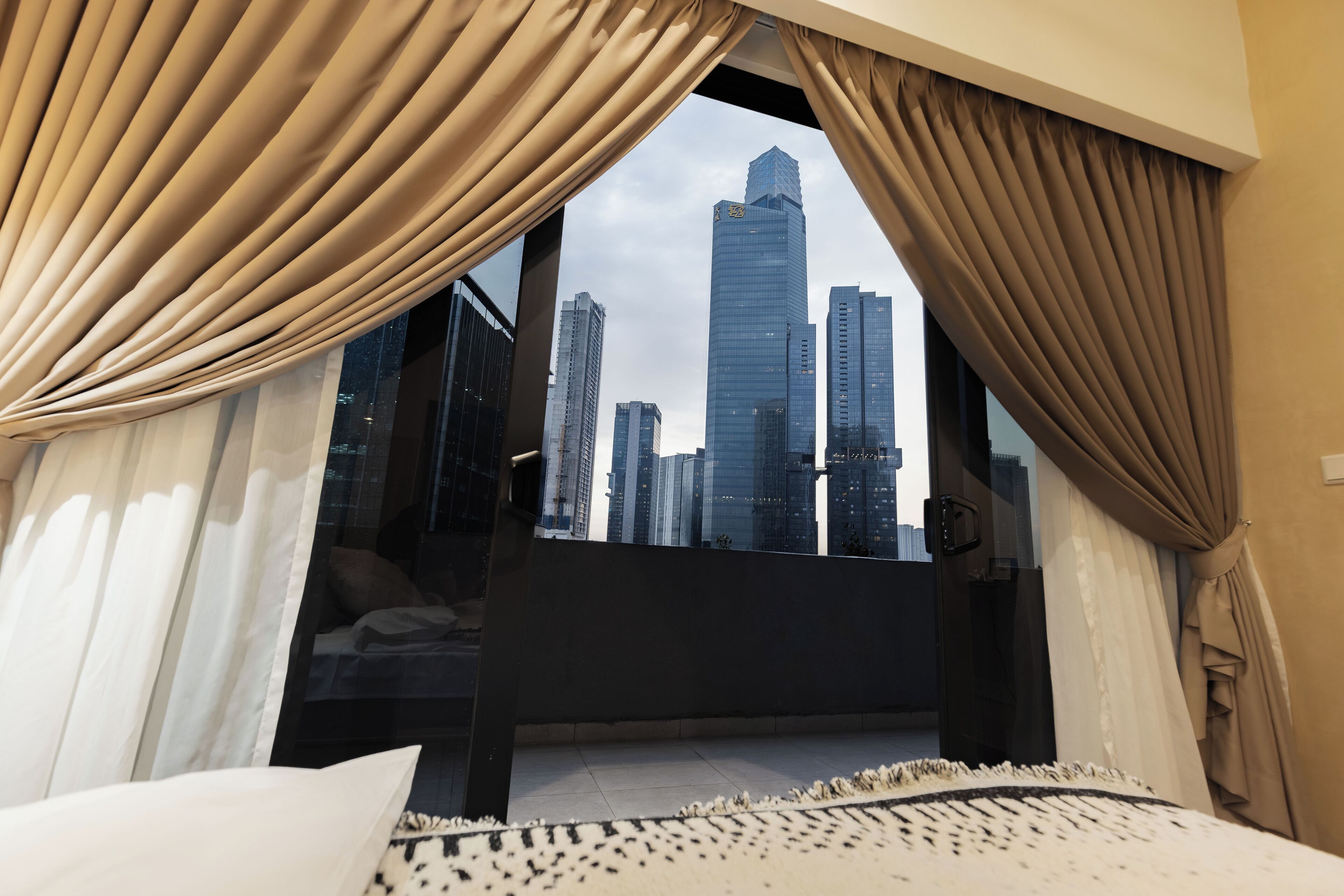 A hotel room with a large window, curtains, a bed, and a view of a city skyline.