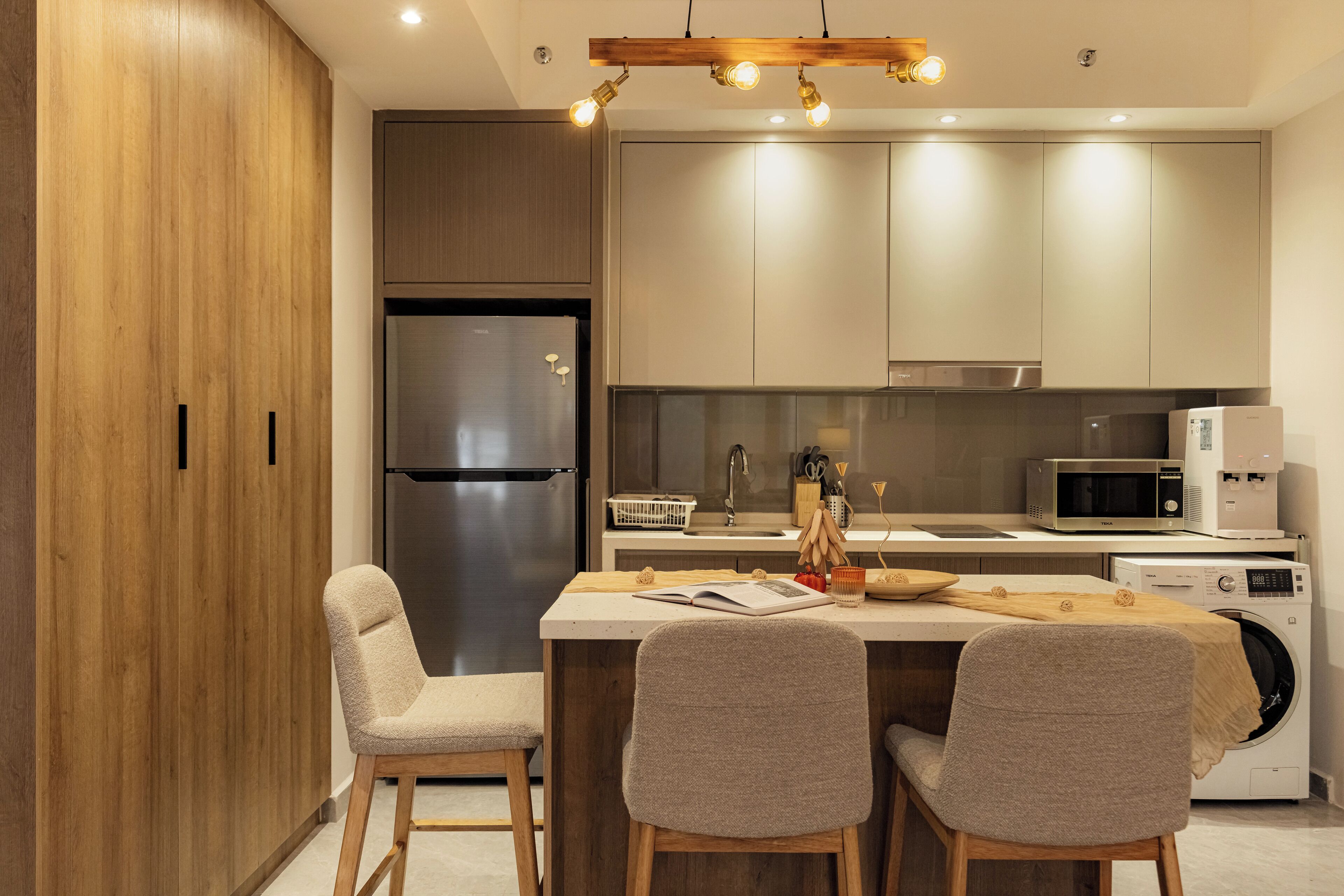 A modern kitchen with a built-in refrigerator, microwave, and washing machine, wooden cabinets, and a dining area with two chairs.