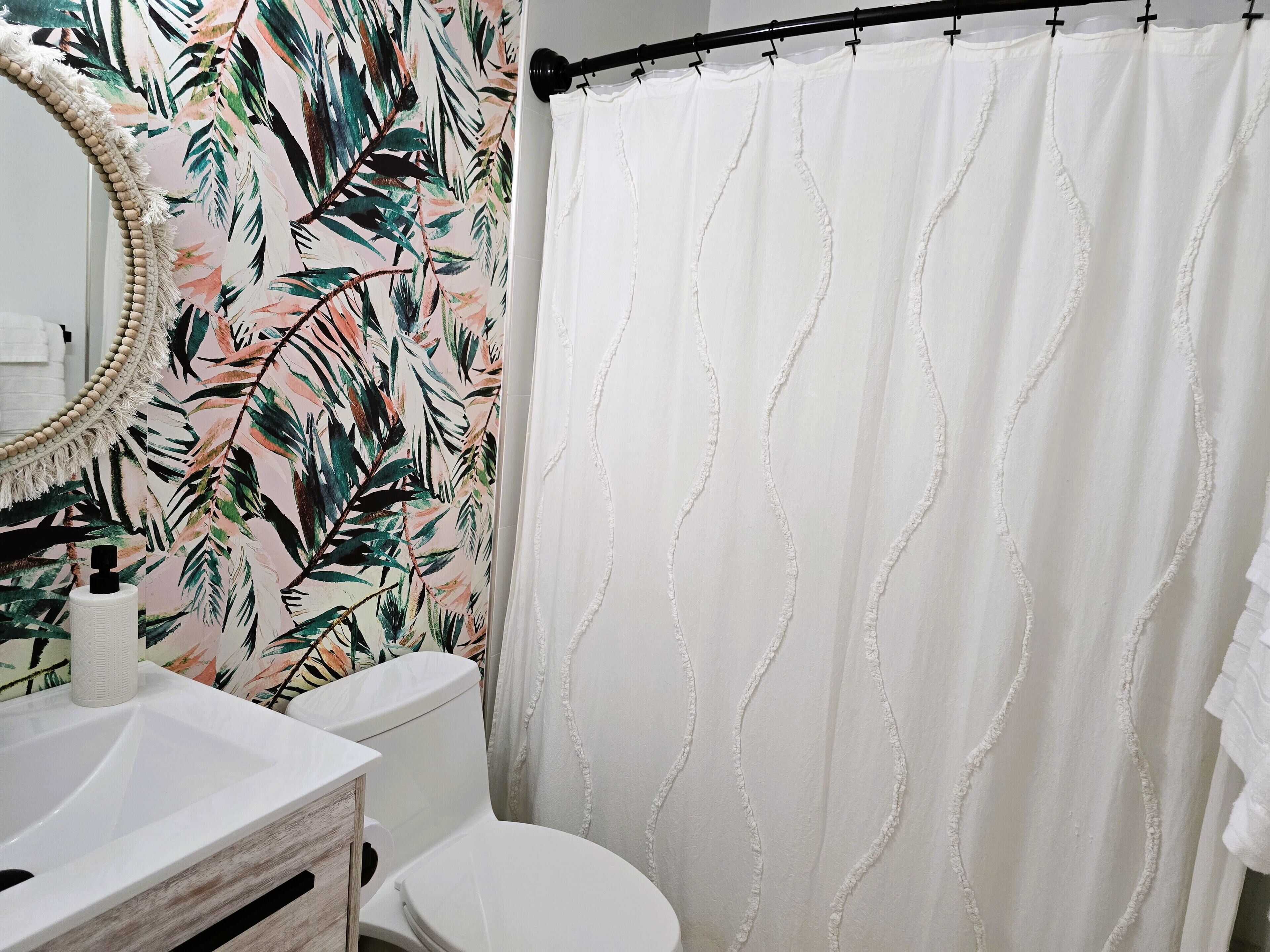 Une salle de bain comprenant des toilettes, un lavabo, un miroir et un rideau de douche orné d’un motif de feuilles.