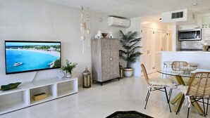 Una sala moderna con un televisor de pantalla plana, un mueble decorativo, una planta en maceta y una zona de comedor con sillas de mimbre.