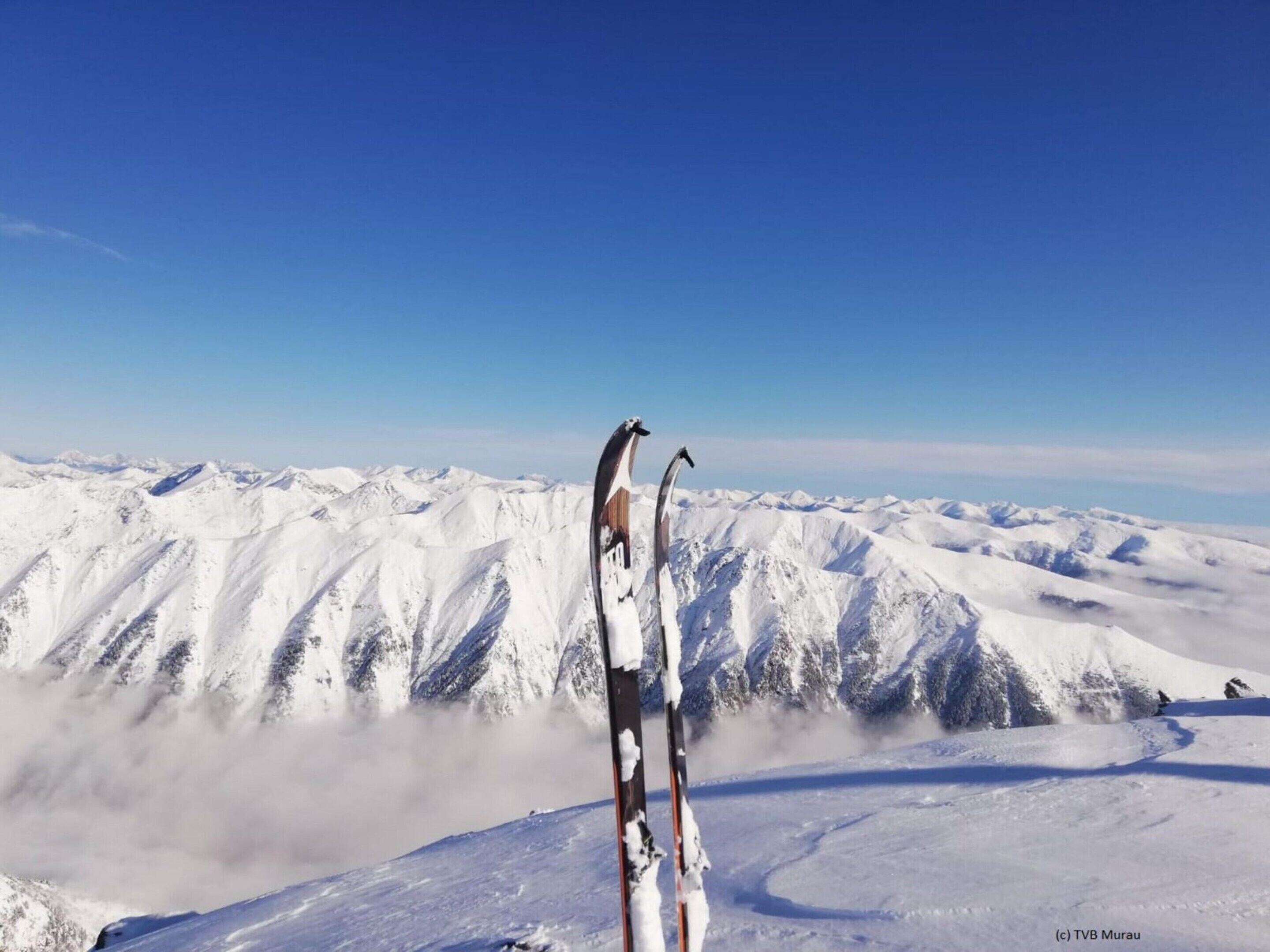 A pair of skis standing upright on a snow-covered mountain peak with a clear blue sky in the background.