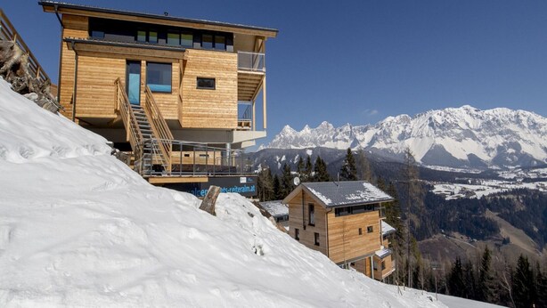 Un moderno chalet in legno con un balcone, sullo sfondo di montagne innevate.