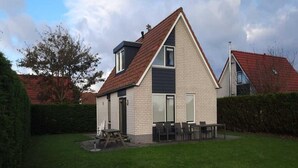 Ein zweigeschossiges Haus mit geneigtem Dach, einer kleinen Garage und einer Terrasse mit Gartenmöbeln.