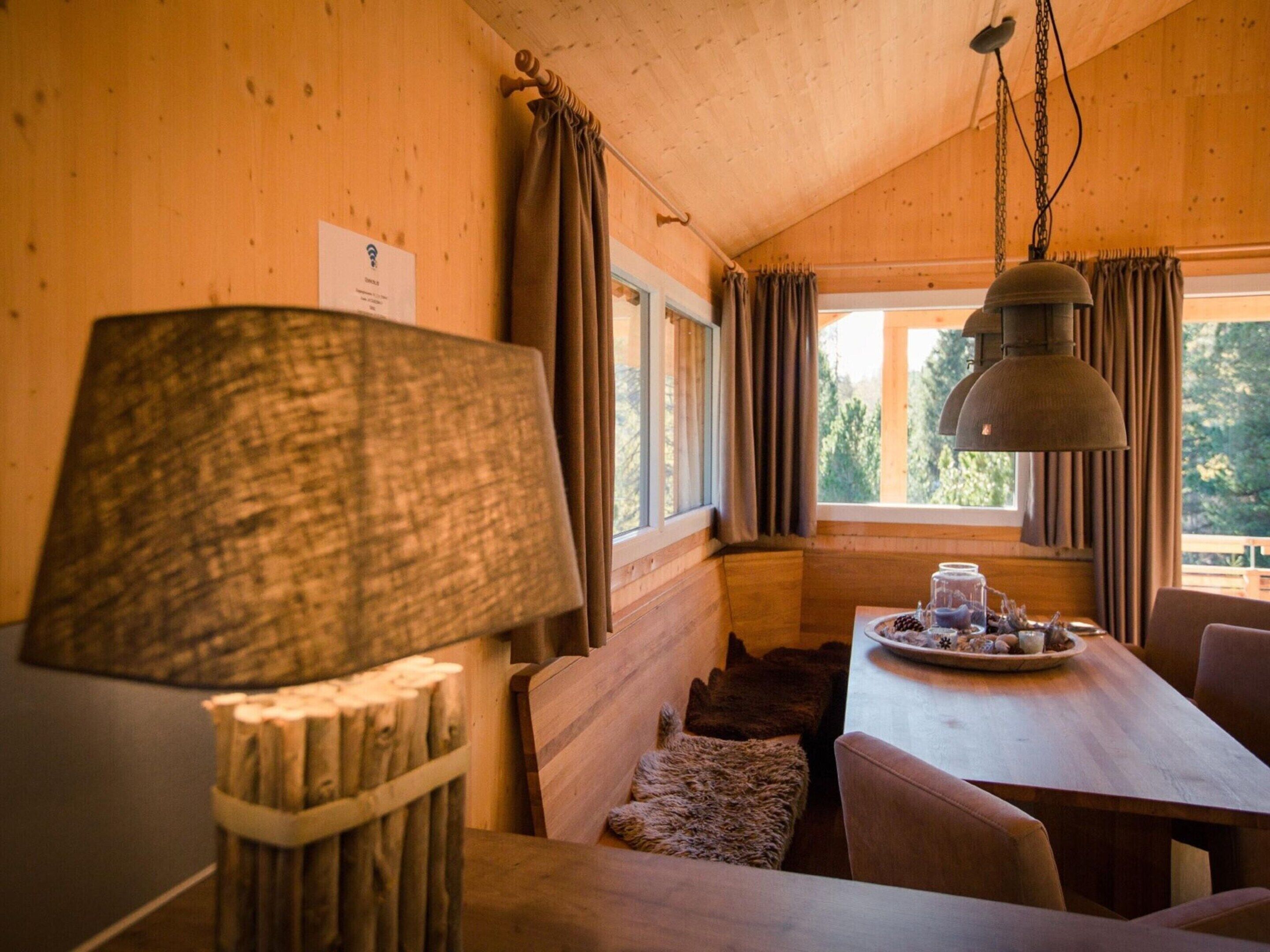 A cozy cabin interior with a wooden table set for dining, a large lamp, and a window with curtains.