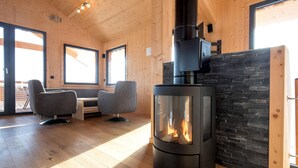 En mysig timmerstuga med en central vedeldad kamin, två grå fåtöljer och ett matbord.