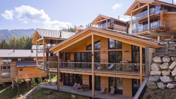 A modern wooden multi-story building with balconies and large windows, surrounded by a mountainous landscape.
