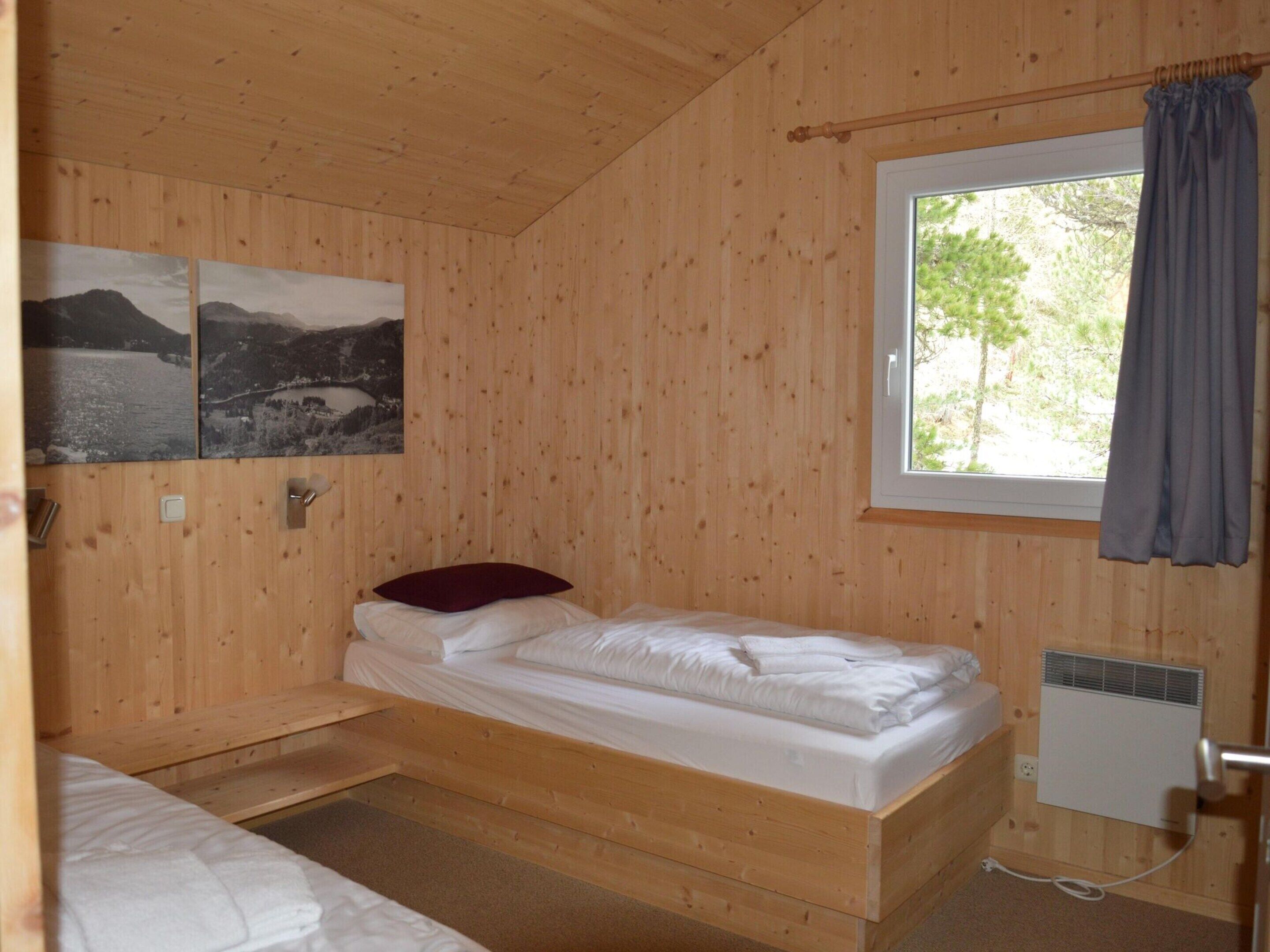 A wooden cabin room with a single bed, a window with curtains, and two black-and-white landscape pictures on the wall.