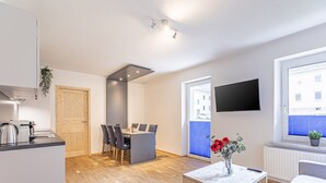Eine moderne Wohnung mit Küche, Essbereich und Wohnzimmer. Sie beinhaltet einen Flachbildfernseher, einen Holztisch und einen Blumentopf.