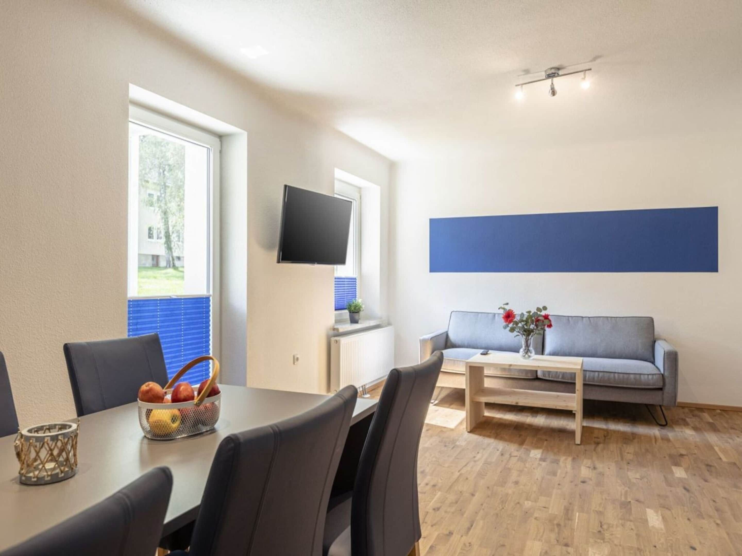 Een moderne kamer met een eettafel, stoelen, een bank, een televisie en een raam met zonwering.