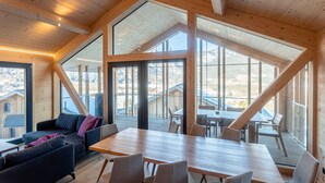 Une pièce spacieuse avec un plafond haut, des poutres en bois, une grande table avec des chaises, un canapé et de grandes fenêtres donnant sur l’extérieur.