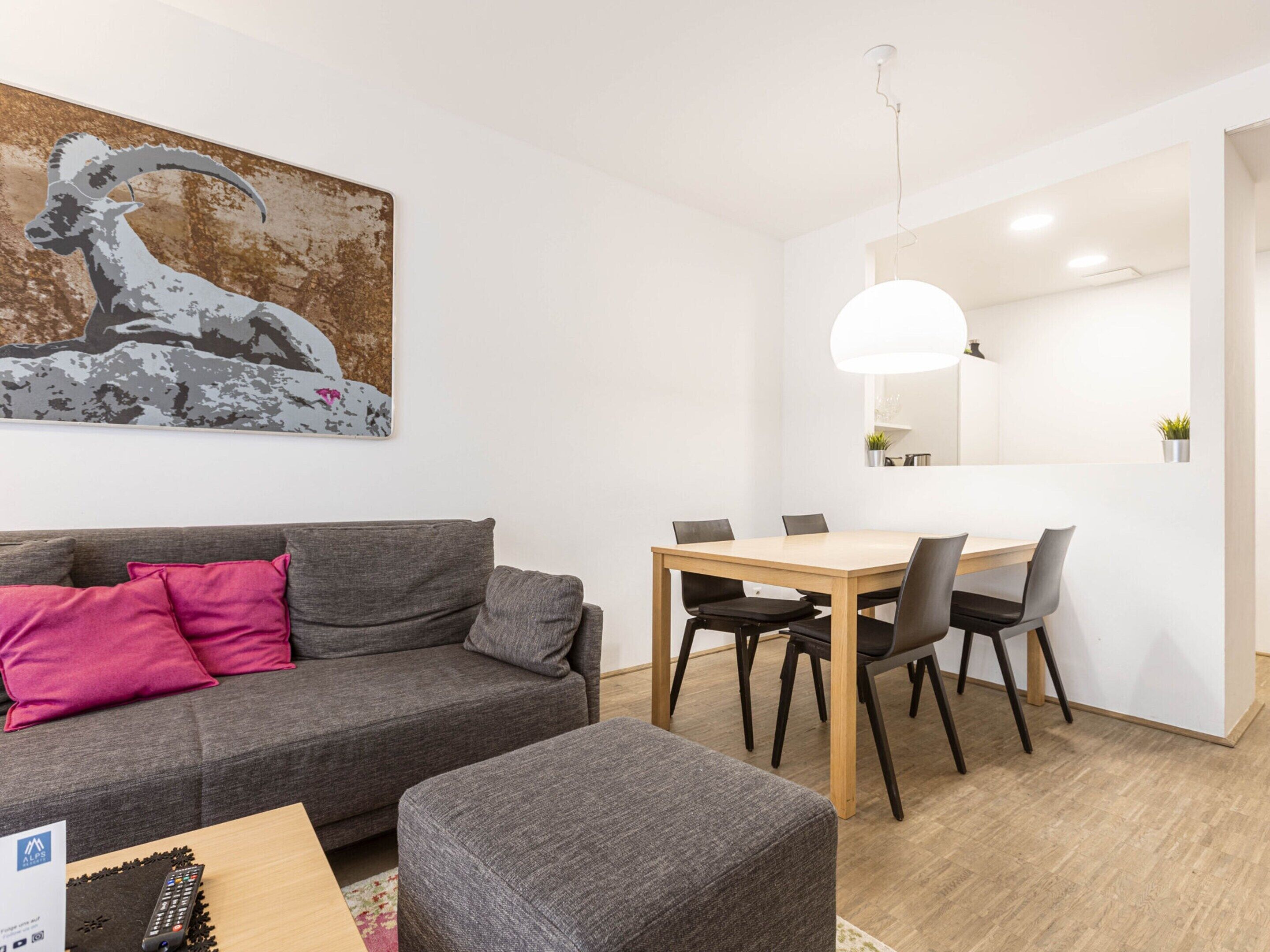 Ett modernt vardagsrum med en grå soffa, ett matbord i trä med stolar, en fjärrkontroll på ett bord och en stor, abstrakt tavla på väggen.