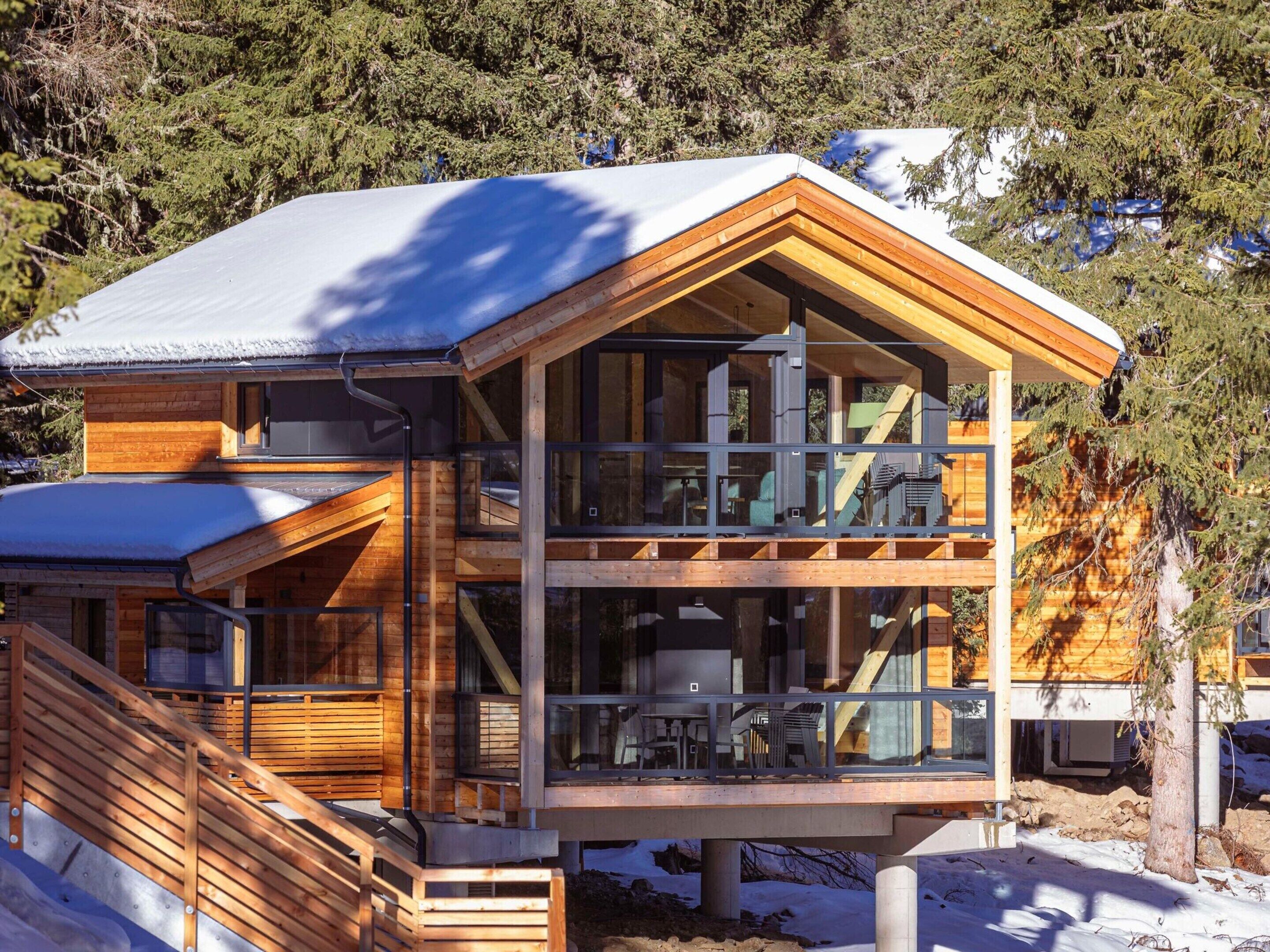 A modern wooden cabin with a balcony, large windows, and a snow-covered roof.