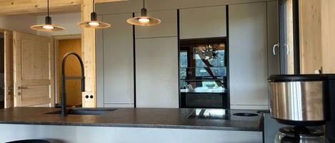 A modern kitchen with wooden ceiling beams, a central island with stools, a coffee machine, and a view of a forest through a window.