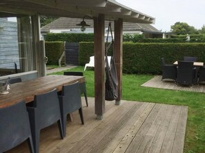 Eine überdachte Terrasse mit Holztable und -stühlen, einer hängenden Tasche und Blick auf einen Hecken und ein Haus im Hintergrund.