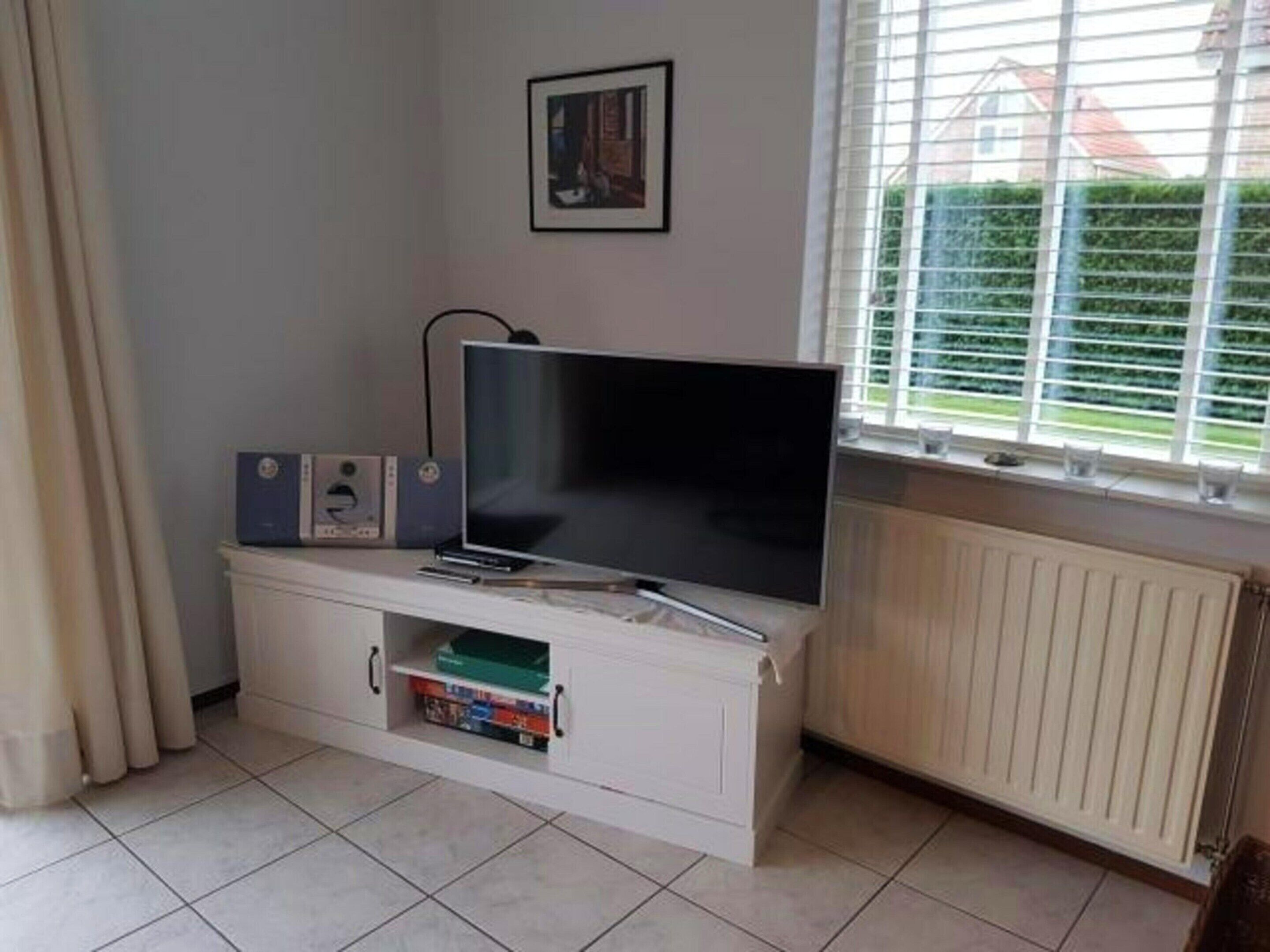 A living room with a flat-screen TV on a white cabinet, a framed picture on the wall, and a window with blinds.