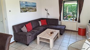 Una sala moderna con un sofá seccional marrón oscuro, una mesa de centro de madera y vistas a la vegetación a través de una ventana con cortinas estampadas.