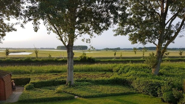 Paisaje rural con césped cuidado, setos y árboles. Se ve una pequeña estructura de madera en primer plano.