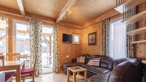 L’intérieur chaleureux d’un chalet avec un plafond en bois, un canapé en cuir, une table à manger avec des chaises et un téléviseur fixé au mur.