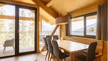 A wooden cabin with a dining area, large windows, and a view of trees and mountains.