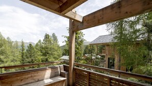 Une terrasse en bois avec un banc et une rambarde, donnant sur une zone boisée où l’on aperçoit un chalet au loin.