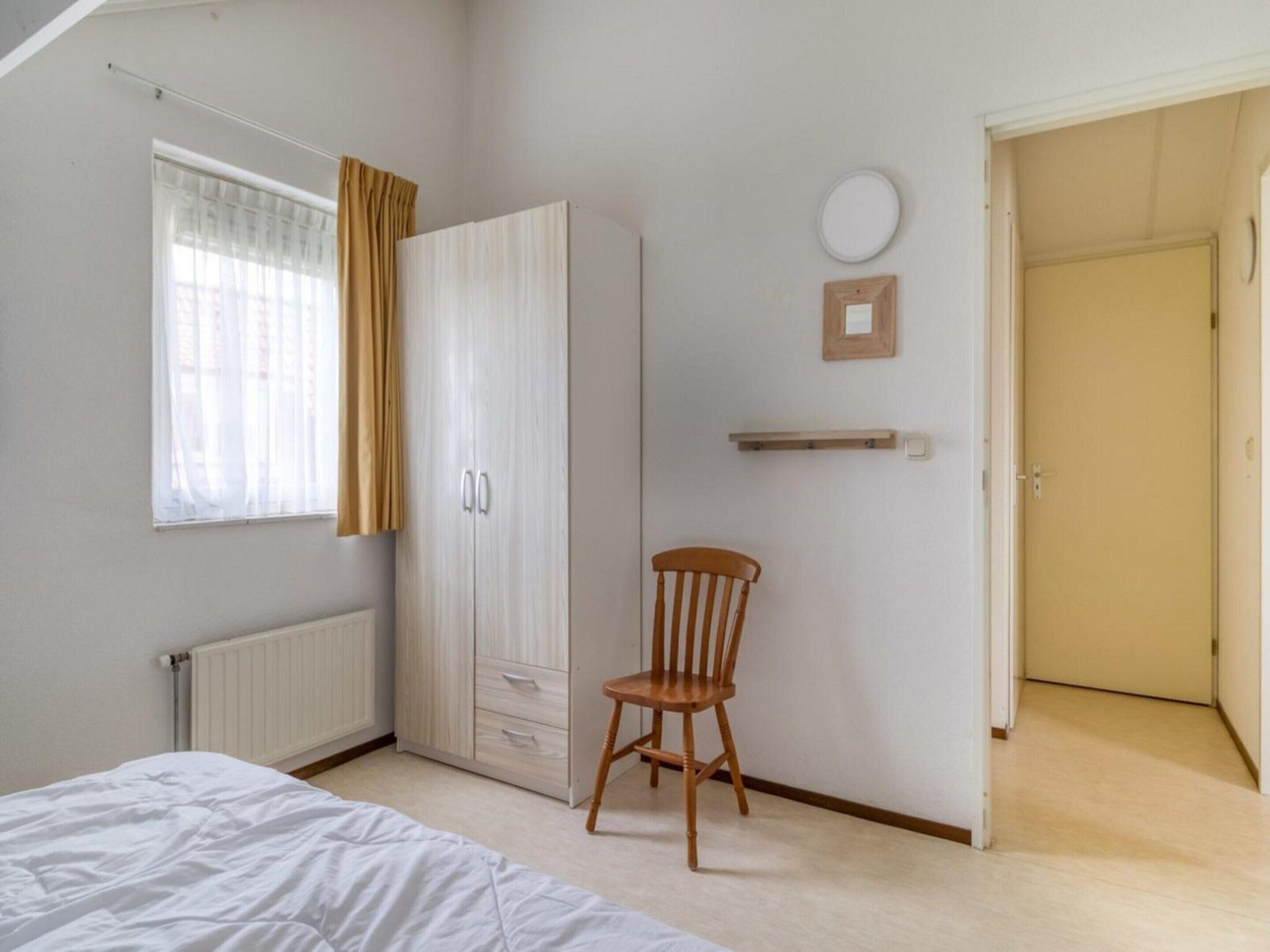 Un dormitorio con cama, armario, silla, radiador, ventana con cortinas, puerta y estante fijado a la pared.
